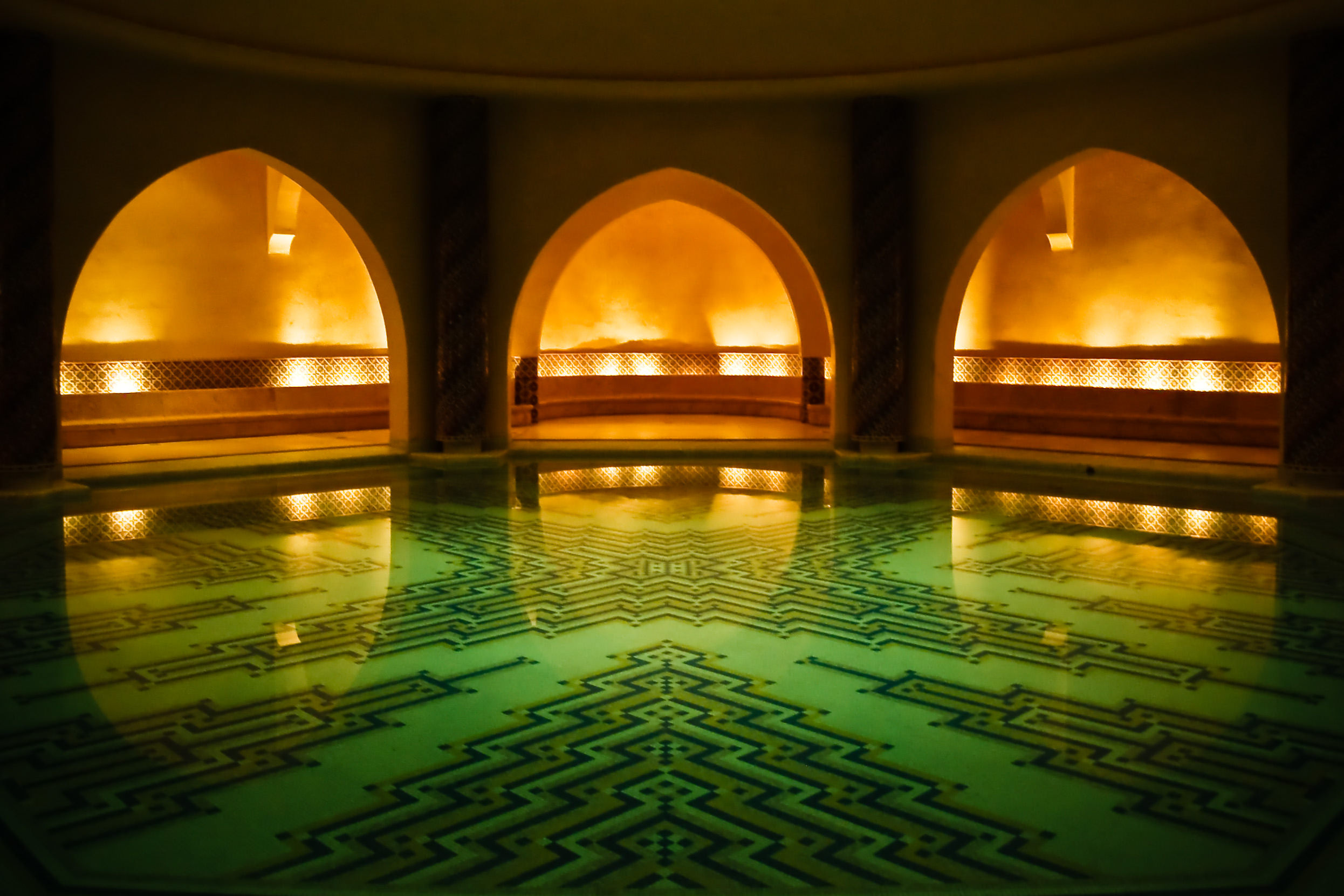 A bath house with a pool in the centre and several arched doorways is lit with low light