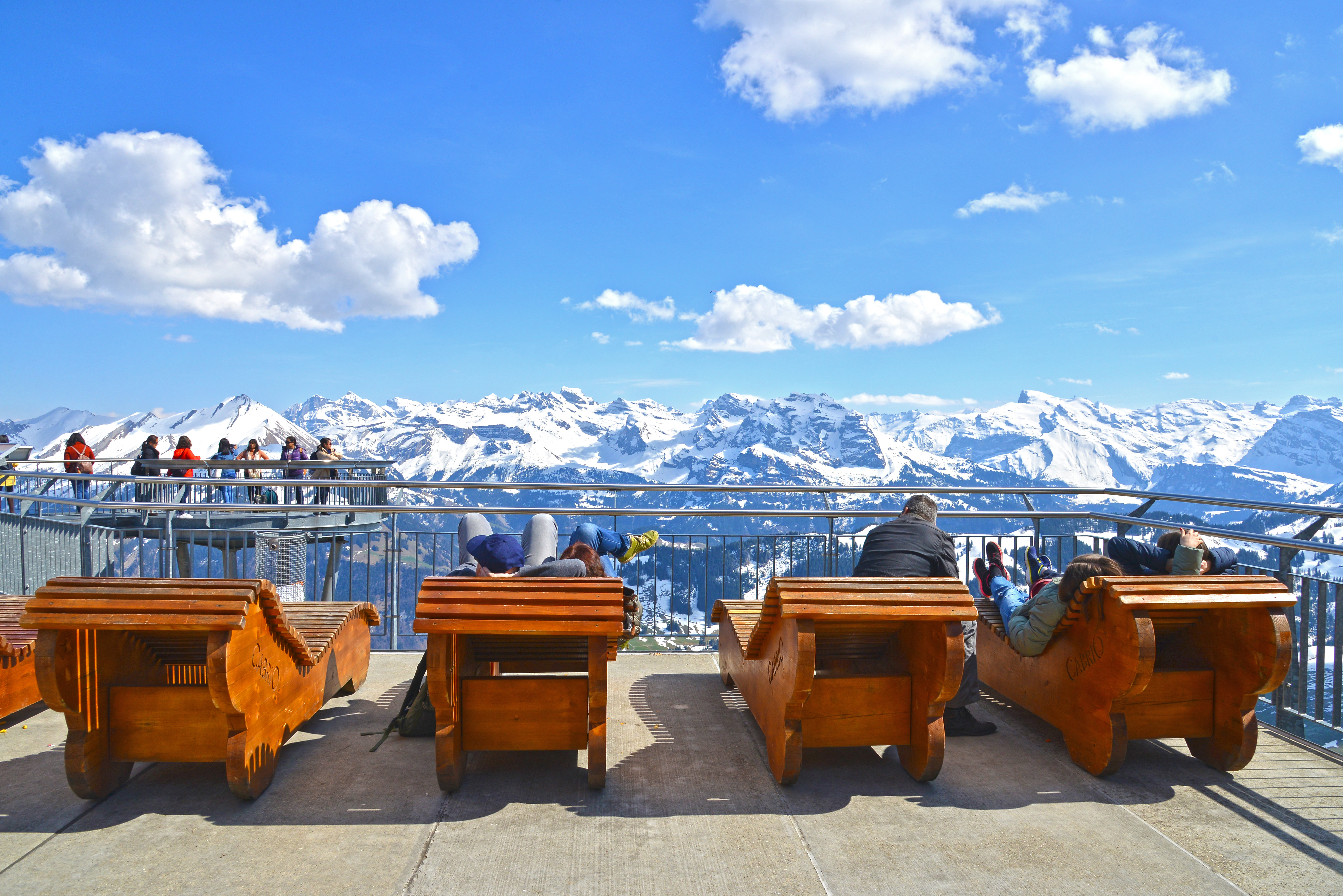 Visitors in wooden deck chairs above snow-capped mountains on the sun terrace atop Stanserhorn Mountain