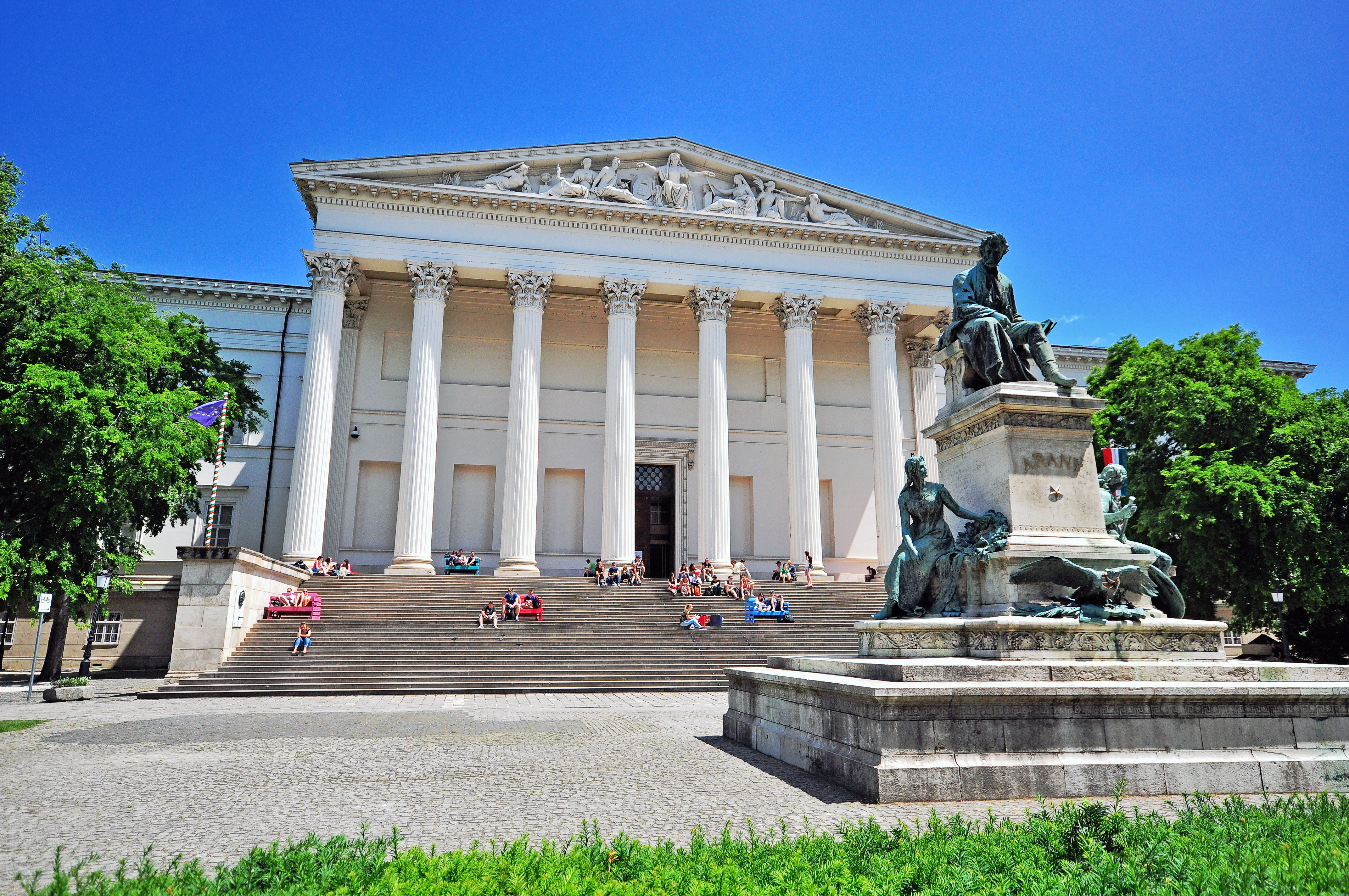 Facade of Hungarian National Museum in Budapest, a large white colonnaded building with people sat on its wide steps