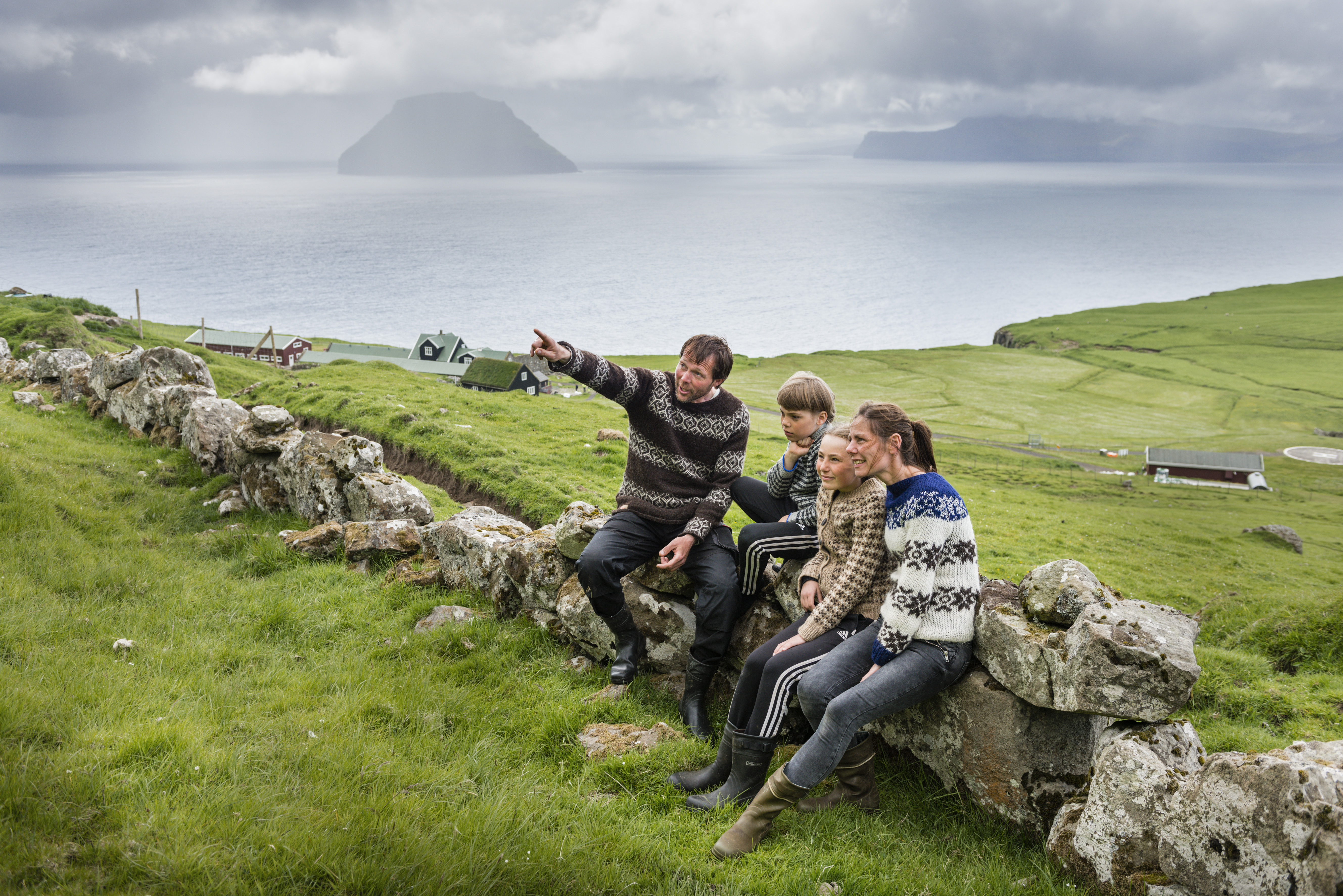 A family sit on a low, rocky wall, all wearing warm woollen clothing and Wellington boots