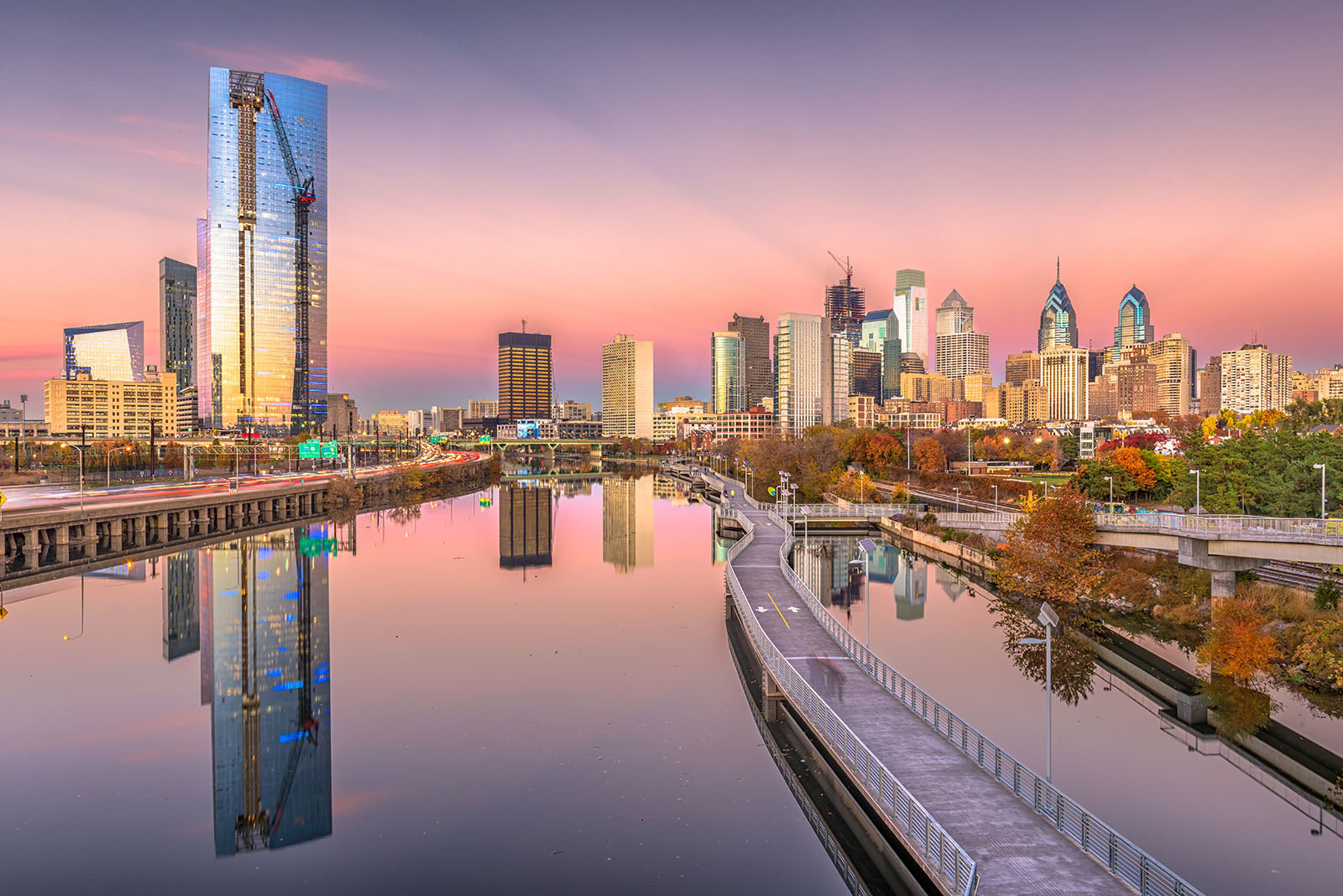 skyline of Philadelphia, with a rose-pink and lavender sunset reflecting off the skyscrapers and the Schuykill River and the boardwalk trail in the foreground