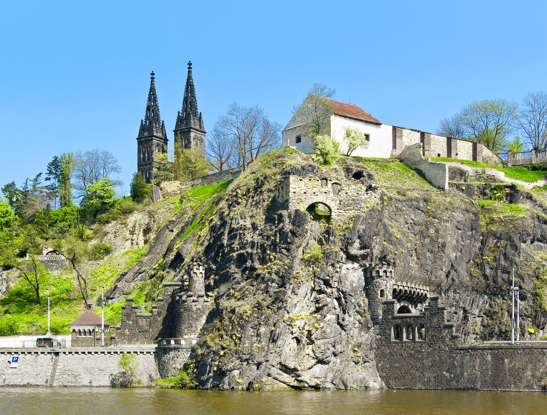 The Vyšehrad Citadel built on the Vltava River; rock ramparts flank the water's edge and rise up the slope to buildings and the twin-spired church.